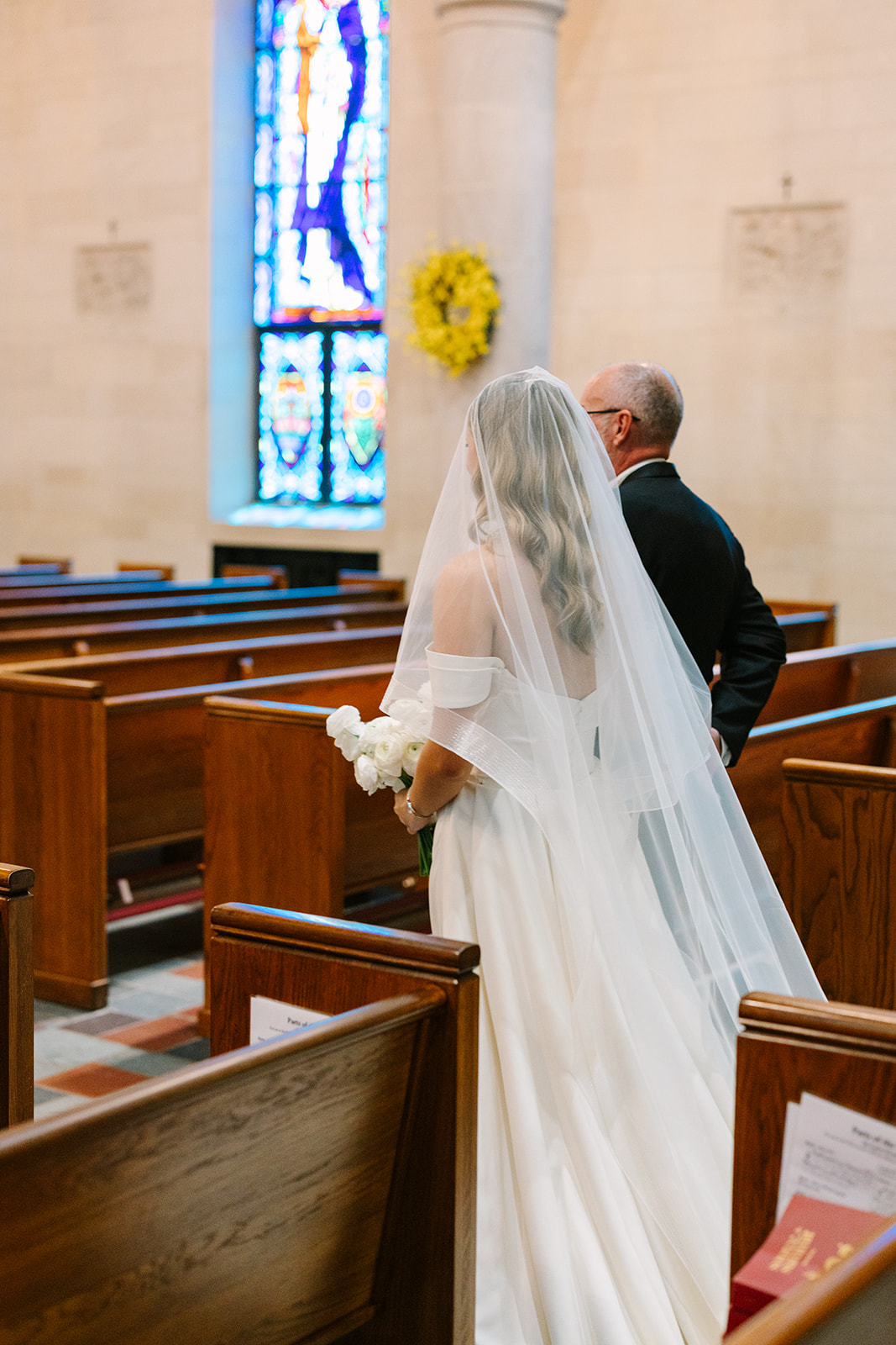 A bride in a white gown and veil walks down the aisle with an older man in a church filled with seated guests, facing the altar.