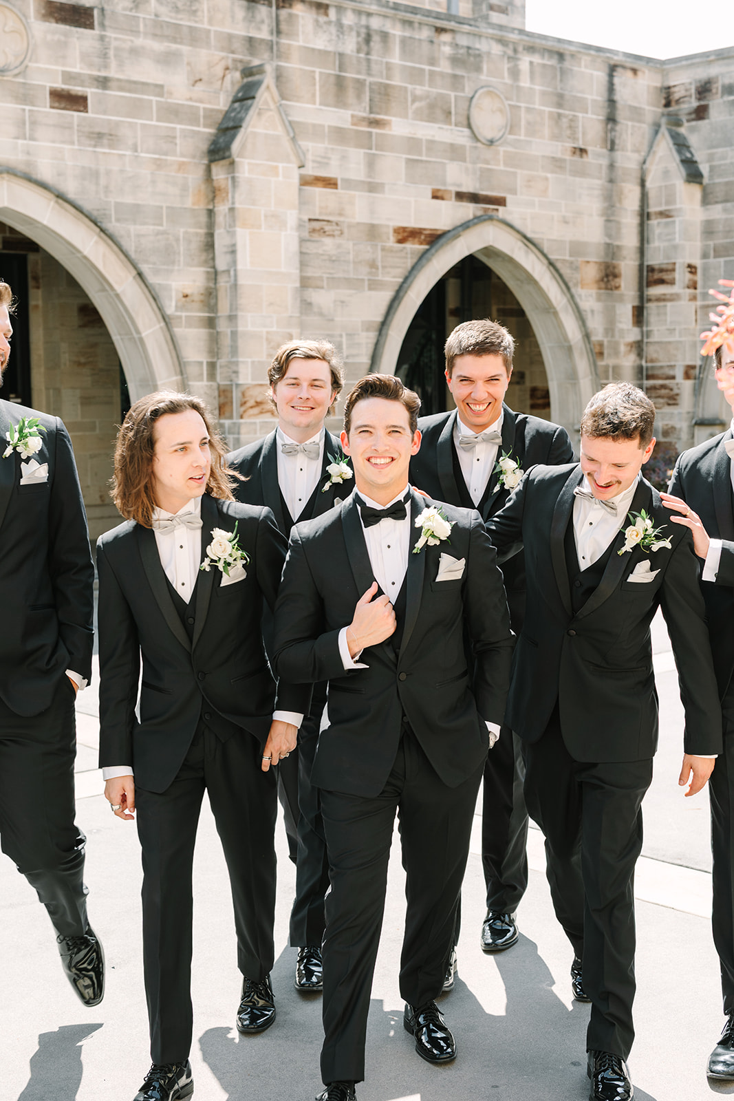 A wedding party poses outdoors in front of a stone building with arched doorways. The group includes the bride, groom, bridesmaids in black dresses, and groomsmen in black suits.