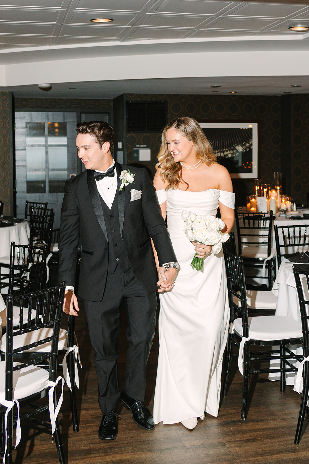 A bride and groom enter a wedding reception hall holding hands, with the bride raising her arms in celebration and guests watching in the background.