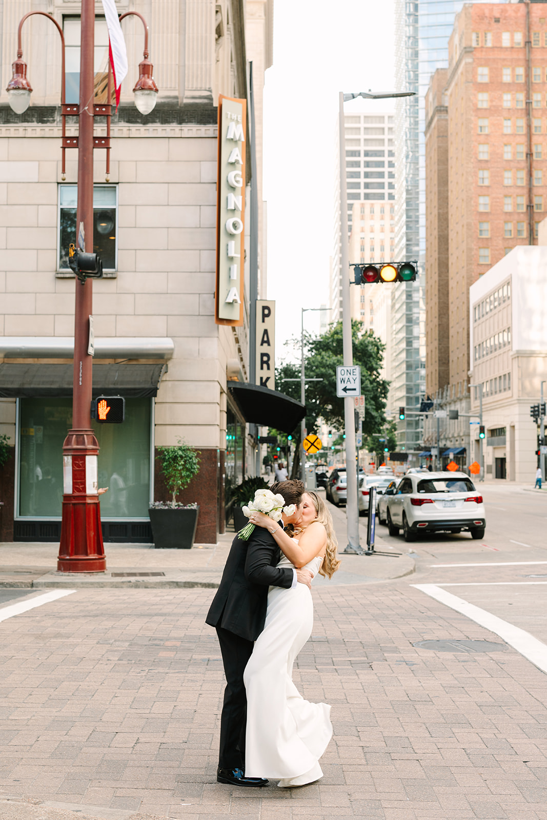 A bride in an off-the-shoulder white gown holds a bouquet of white flowers and stands beside a groom in a black tuxedo for a wedding at the Magnolia Hotel in Houston