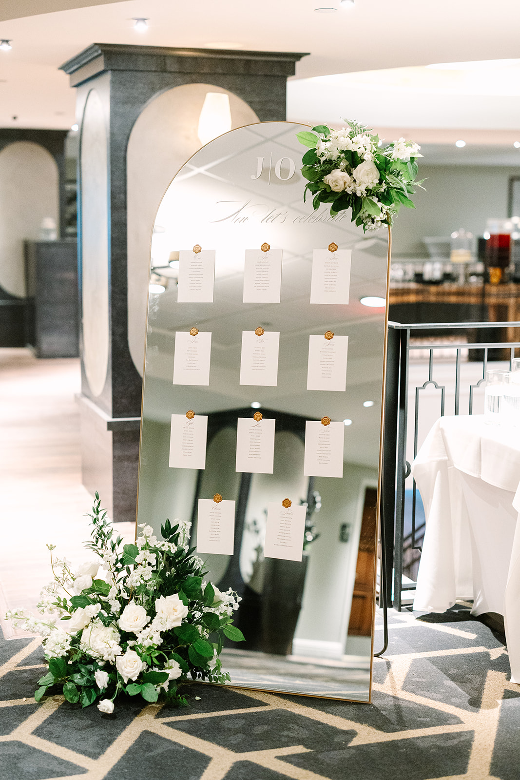 A large mirrored seating chart display with white cards, wax seals, and floral arrangements at the top and bottom, set at Magnolia Hotel in Houston