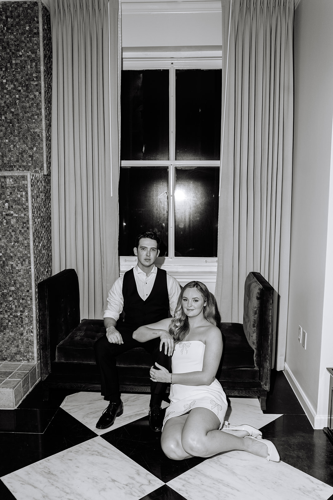 A man in a vest sits on a bench while a woman in a strapless dress sits on the floor beside him on a checkered tile floor in front of a window with closed curtains.