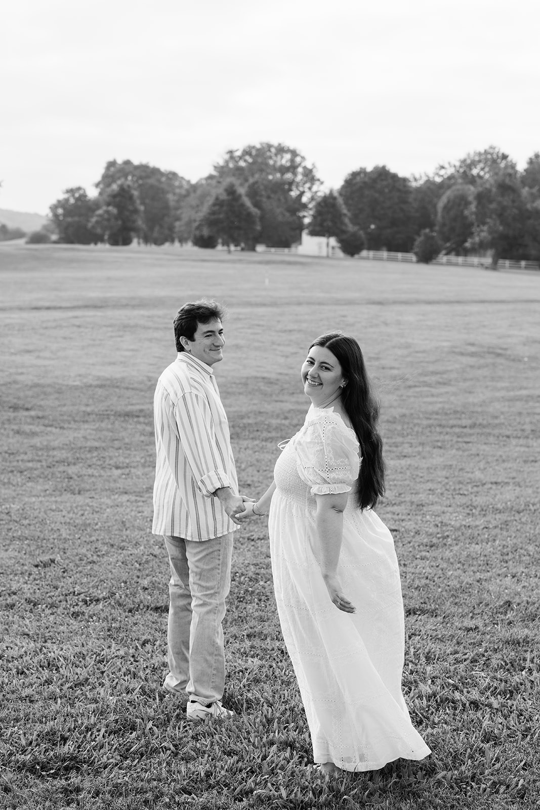 A couple walks across a grassy field with a large tree and distant hills in the background under a bright, overcast sky for destination engagement photos