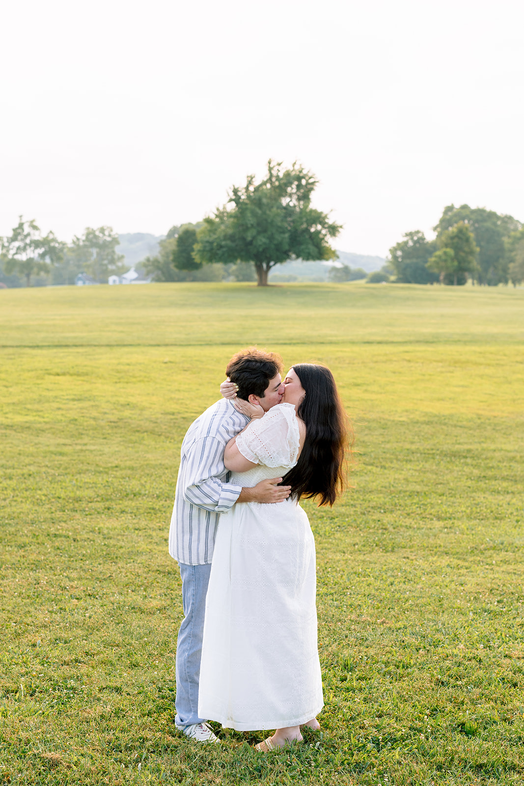 A couple walks across a grassy field with a large tree and distant hills in the background under a bright, overcast sky for destination engagement photos