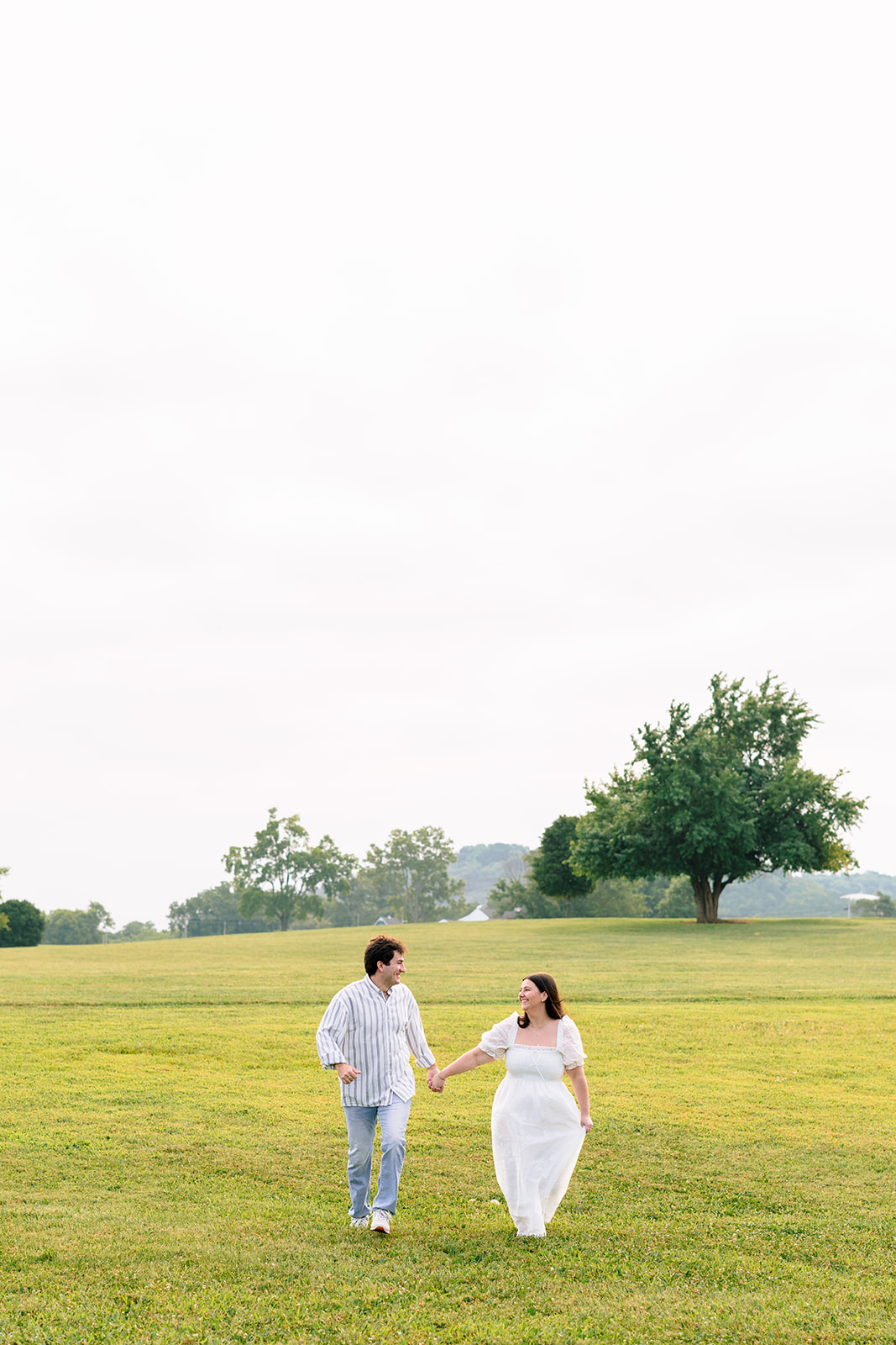 A couple holding hands walks across a grassy field with a large tree and distant hills in the background under a bright, overcast sky for destination engagement photos