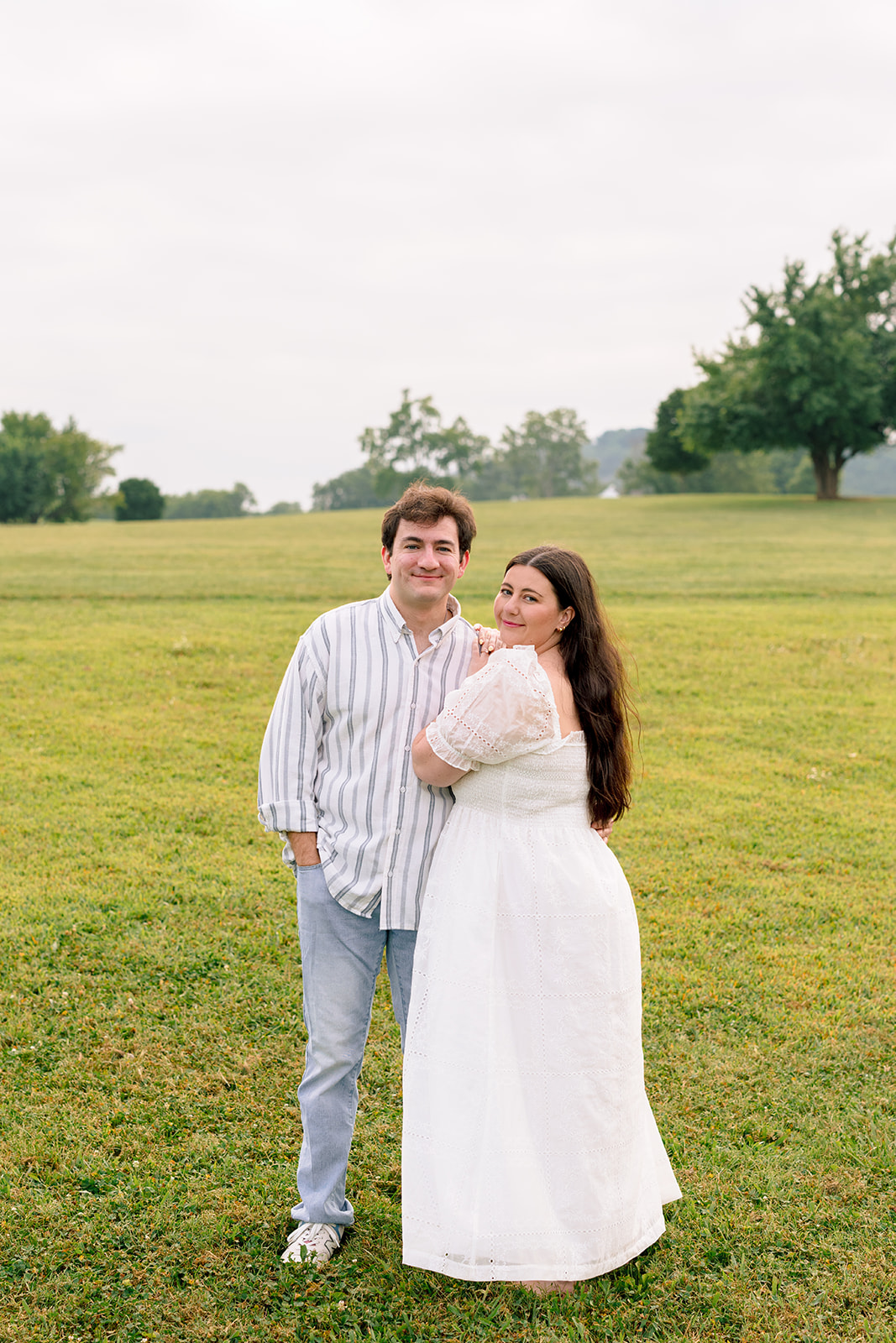 A couple walks across a grassy field with a large tree and distant hills in the background under a bright, overcast sky 