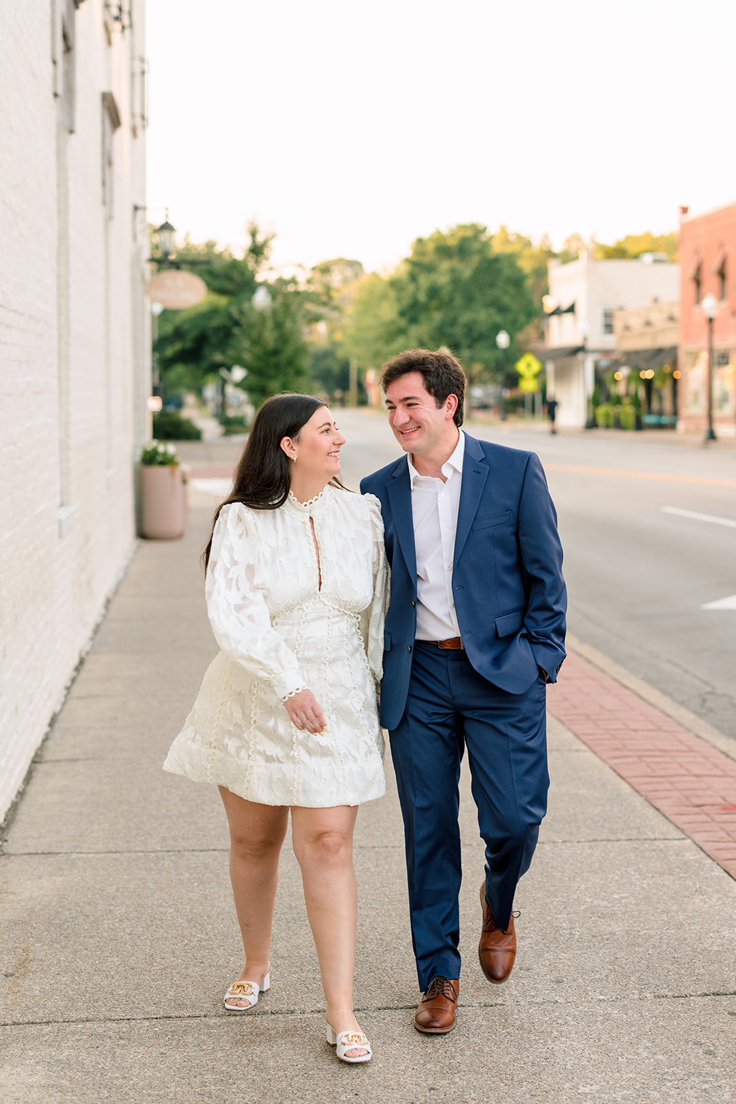  A man in a blue suit and a woman in a white dress hold hands while walking outside on a paved street, with trees and buildings in the background. 