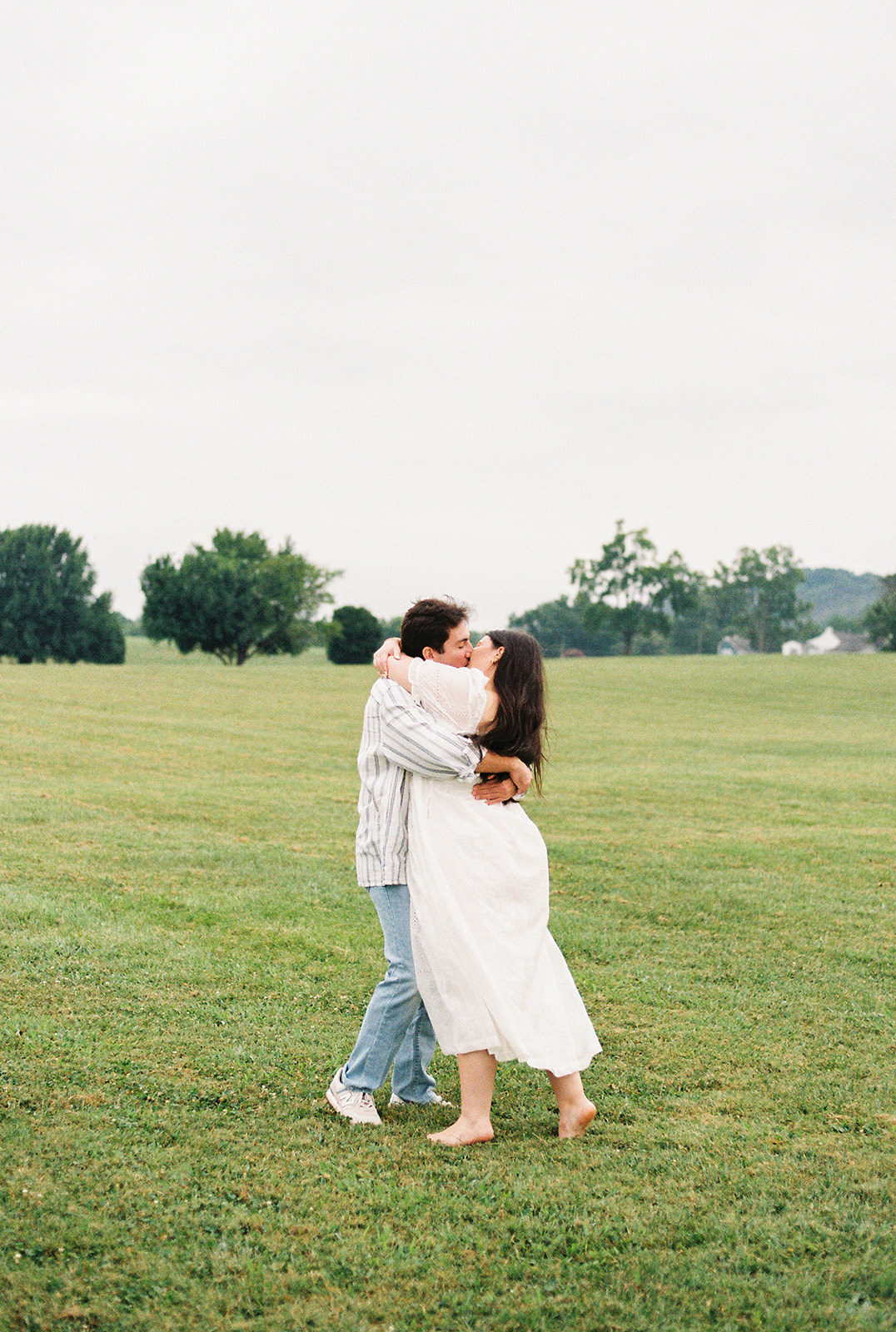 A couple walks across a grassy field with a large tree and distant hills in the background under a bright, overcast sky for destination engagement photos