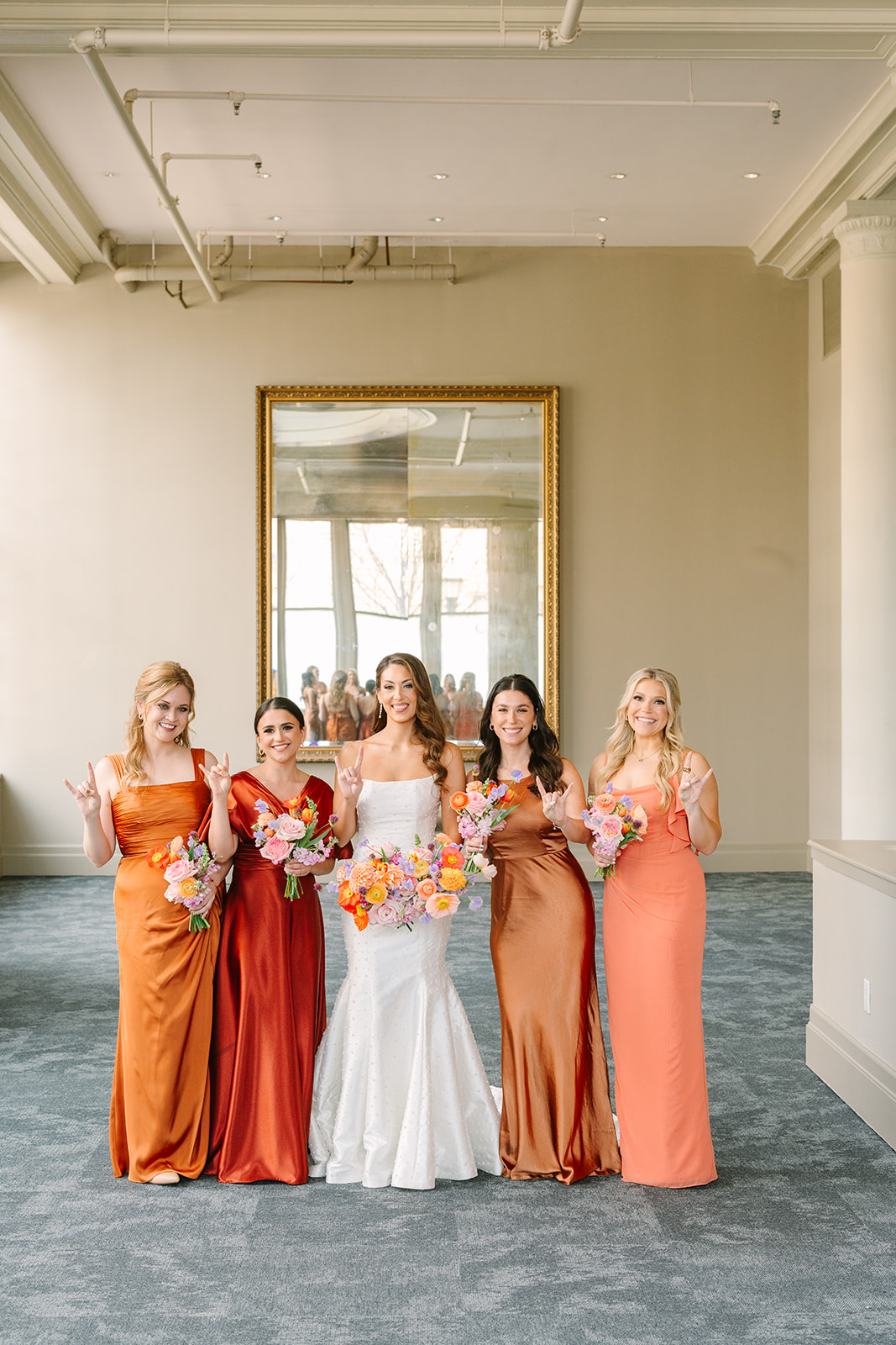 A bride in a white gown stands with nine bridesmaids in coordinating burnt orange dresses, all holding colorful bouquets, in a bright room with large windows.