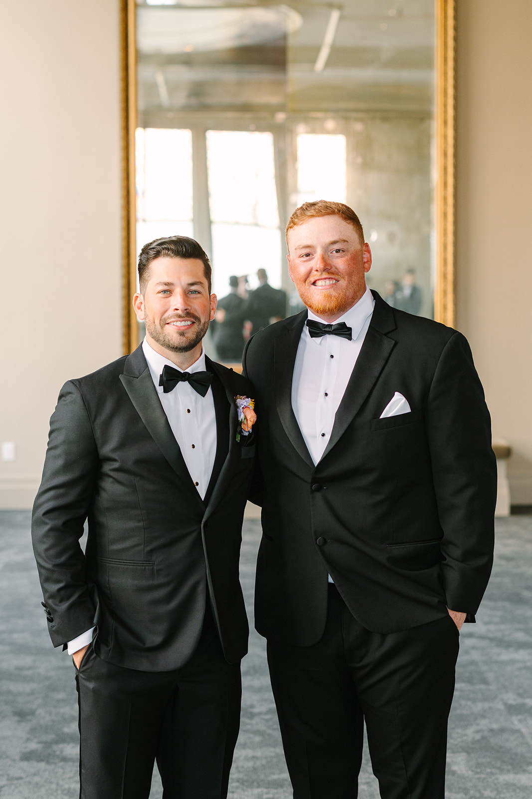 A group of men in black tuxedos stand together indoors, with one man at the center holding his jacket lapel. A large mirror is visible on the wall behind them.