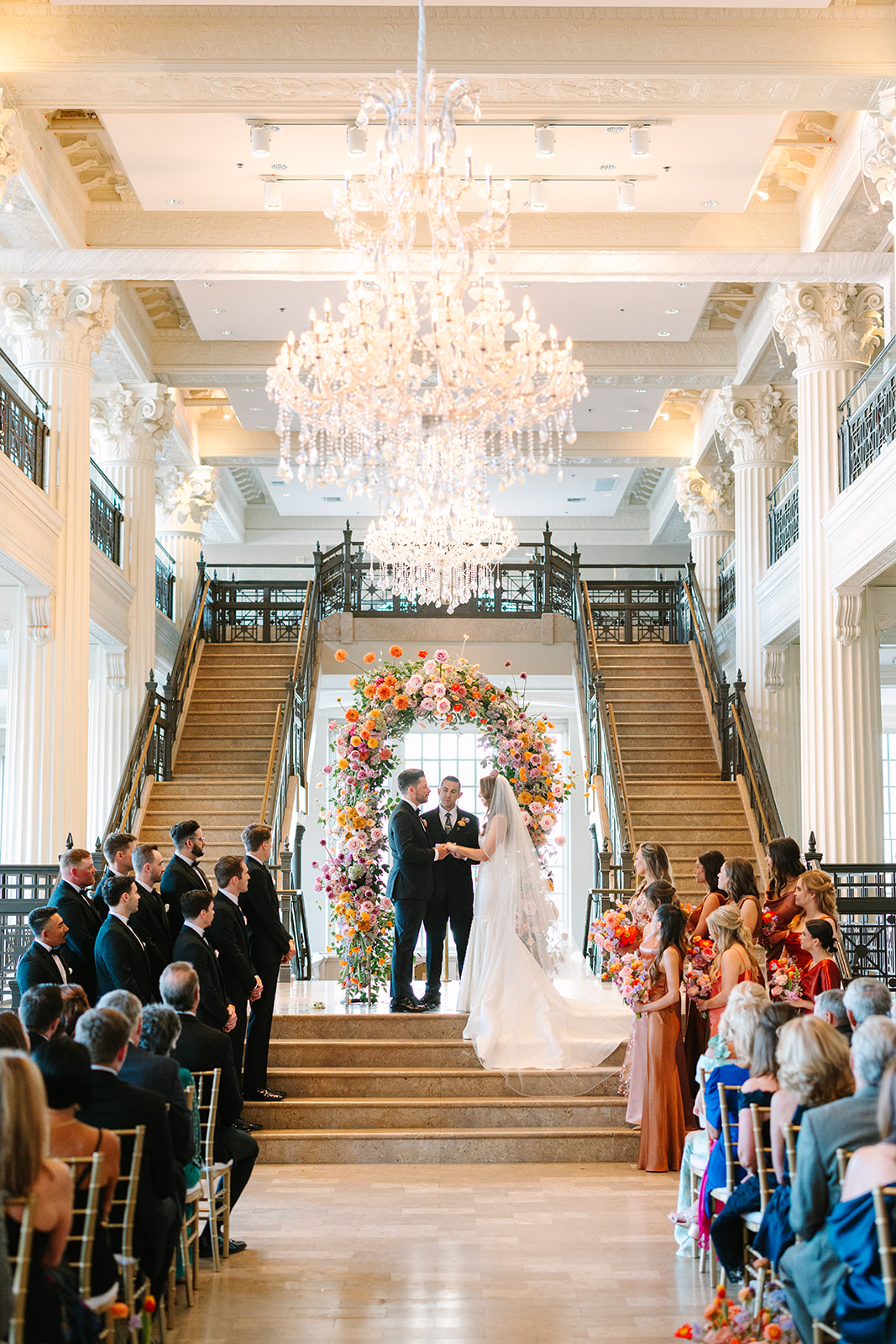 A couple stands with an officiant under a floral arch at the top of a grand staircase, surrounded by guests in a large, elegant hall with chandeliers at the corinthian houston