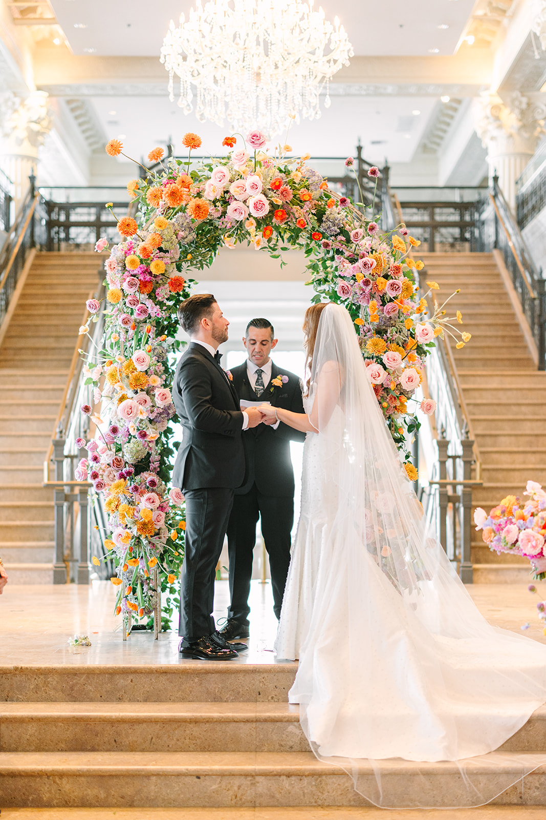 A couple stands with an officiant under a floral arch at the top of a grand staircase, surrounded by guests in a large, elegant hall with chandeliers at the corinthian houston