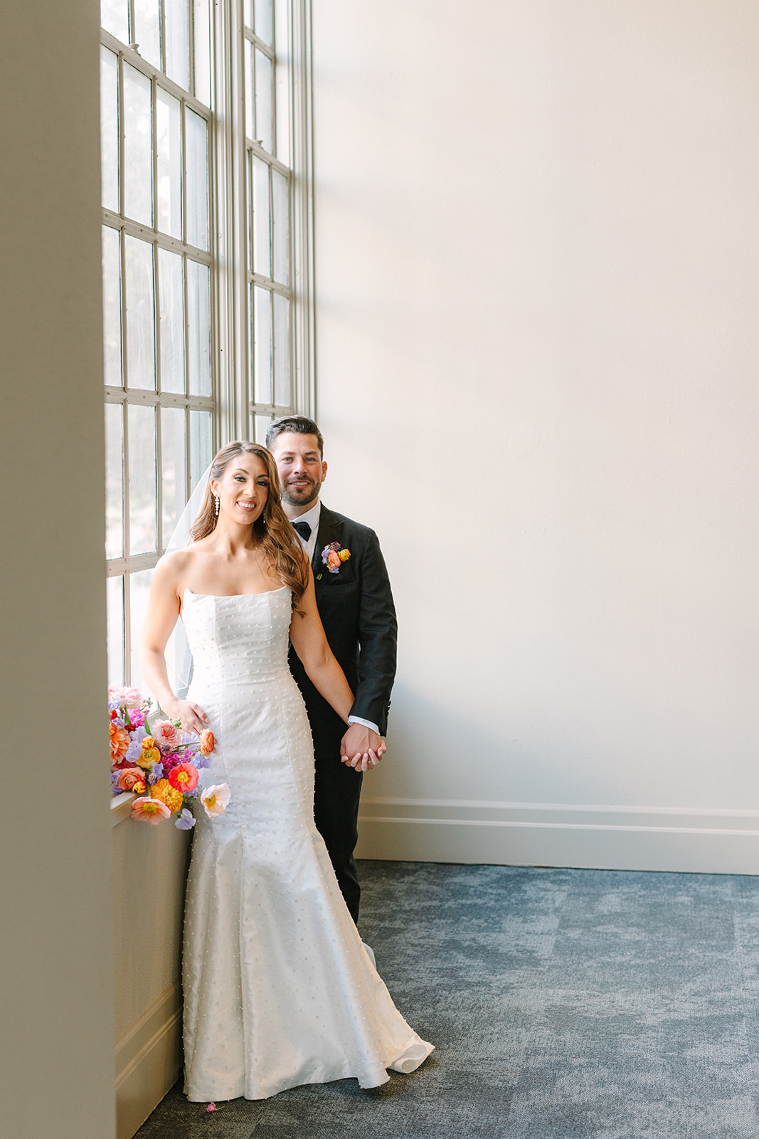 A bride in a white gown holding a colorful bouquet walks alongside a groom in a black tuxedo down a hallway with large windows for a wedding at the corinthian houston