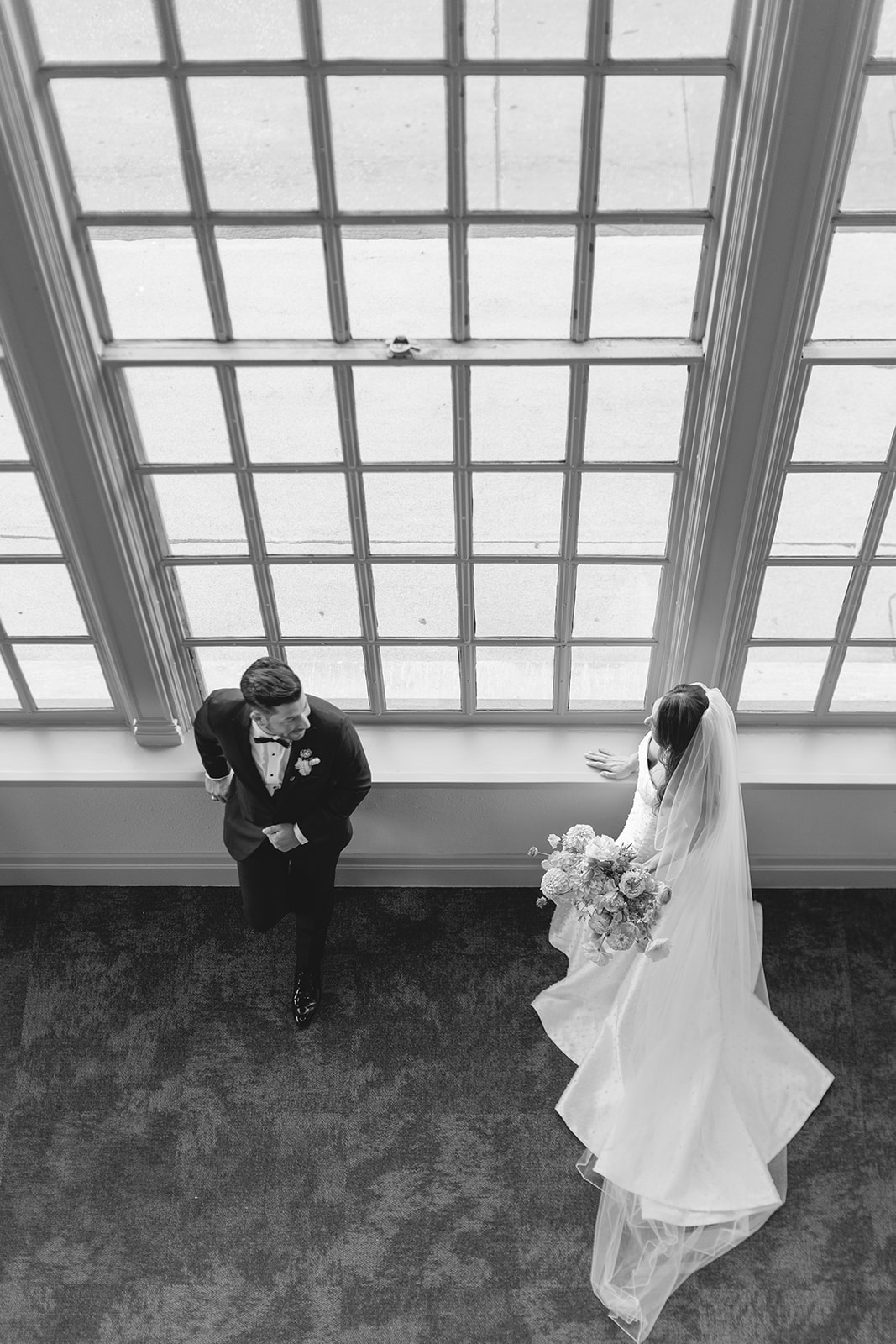 A bride in a white gown holding a colorful bouquet walks alongside a groom in a black tuxedo down a hallway with large windows for a wedding at the corinthian houston