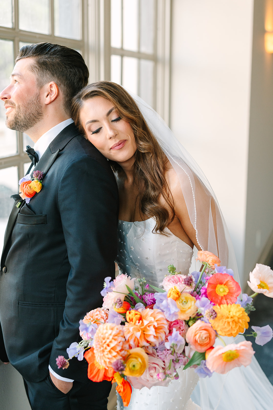 A bride in a white gown holding a colorful bouquet walks alongside a groom in a black tuxedo down a hallway with large windows for a wedding at the corinthian houston