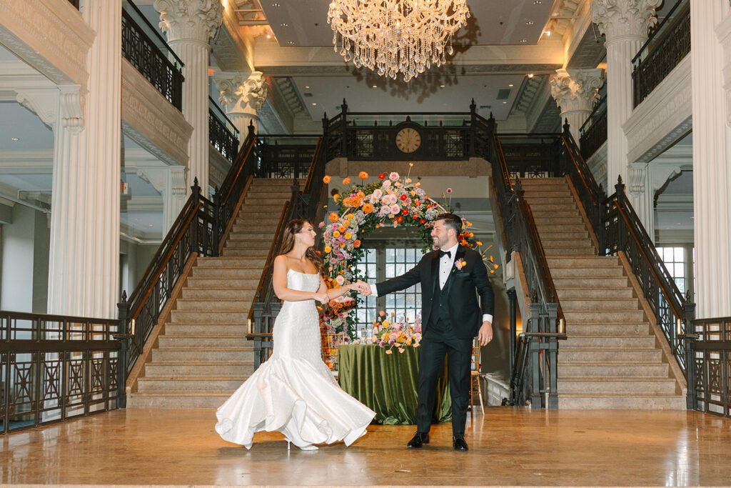 A bride and groom hold hands and dance in an elegant hall with grand staircases, a chandelier, and a floral arrangement behind them.