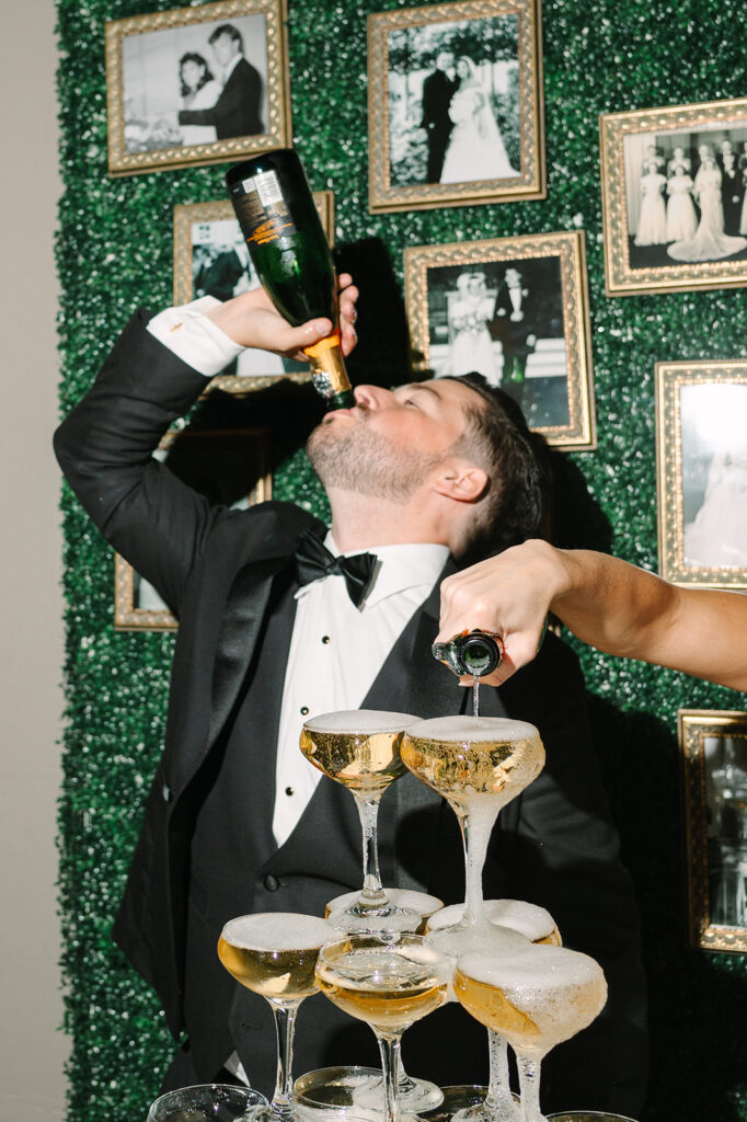 A bride and groom pour champagne into a tower of glasses at their wedding reception, with a cake and floral centerpiece on the table and framed photos on the wall behind them.