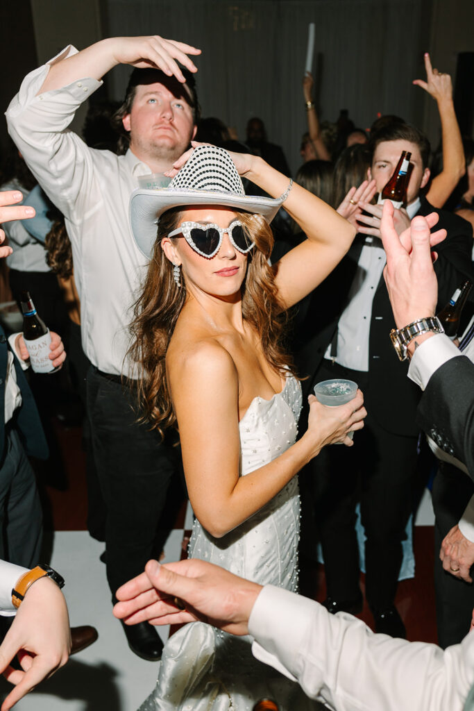 A group of people dance energetically at a formal event; one woman in a white dress holds a drink and raises her arm, while a man in a suit dances barefoot.