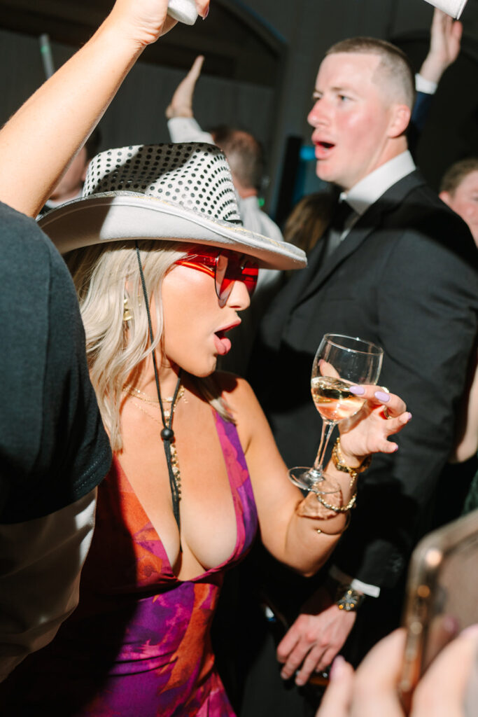 A group of people dance energetically at a formal event; one woman in a white dress holds a drink and raises her arm, while a man in a suit dances barefoot.