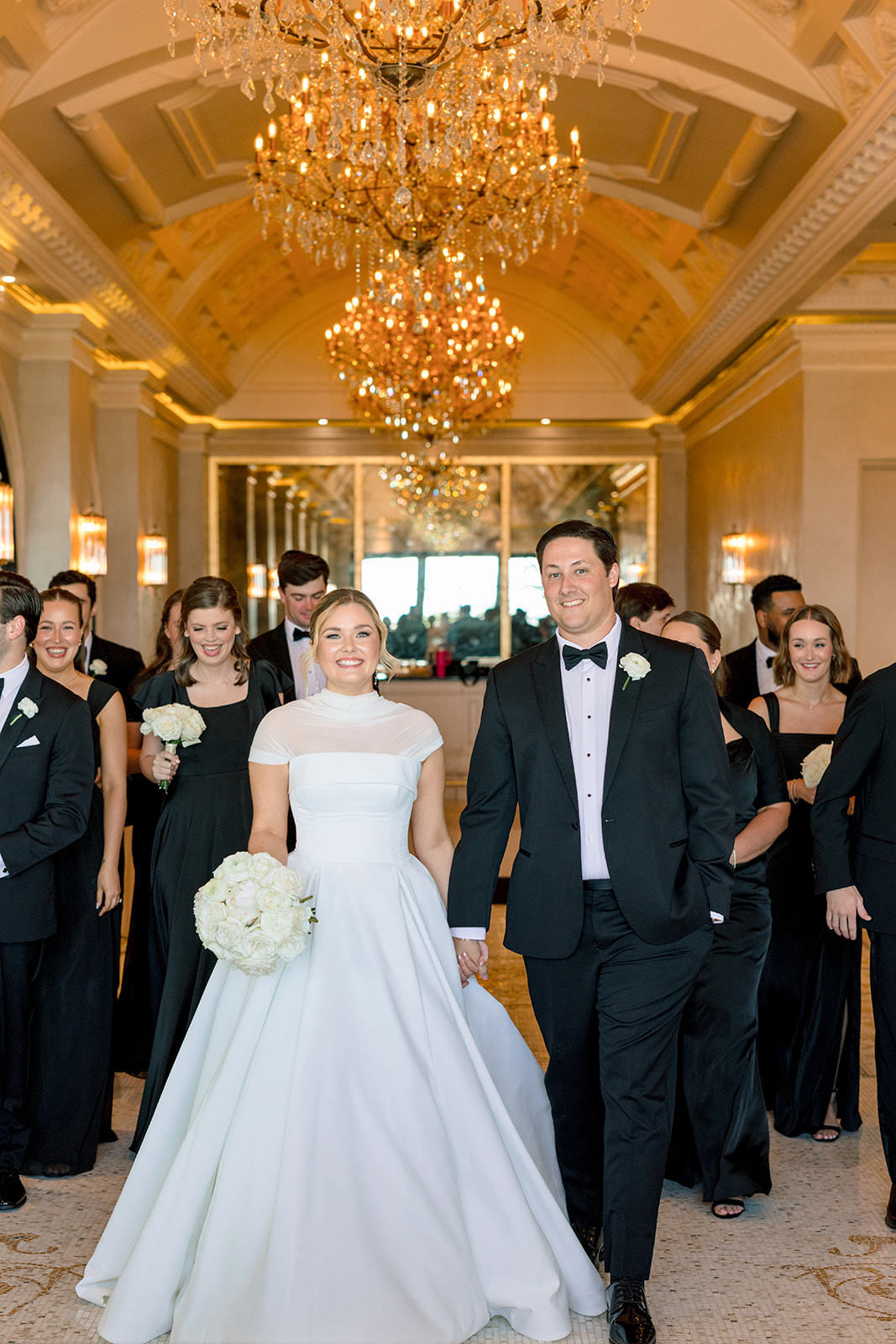 A bride in a white dress stands with ten bridesmaids in black dresses, all holding white bouquets, under ornate chandeliers in an elegant hallway for a Le Tesserae Wedding in Houston
