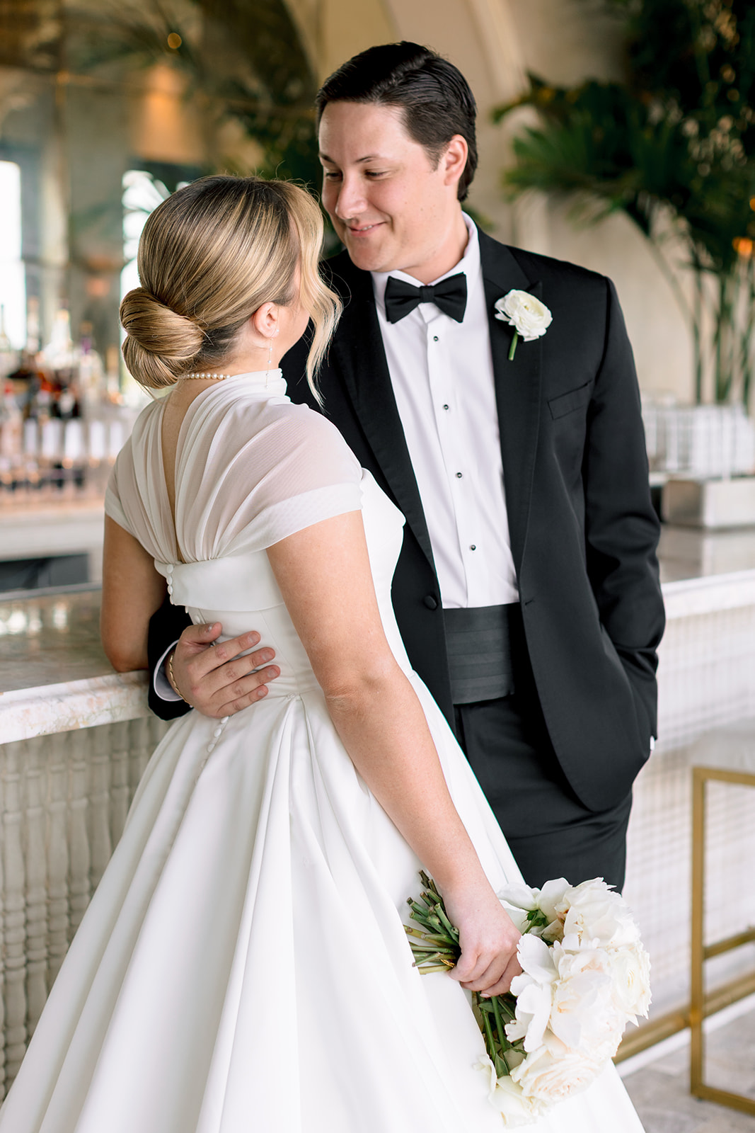 A bride and groom stand together in an elegant room with large windows; the bride wears a long white gown and holds a bouquet for a Le Tesserae Wedding in Houston