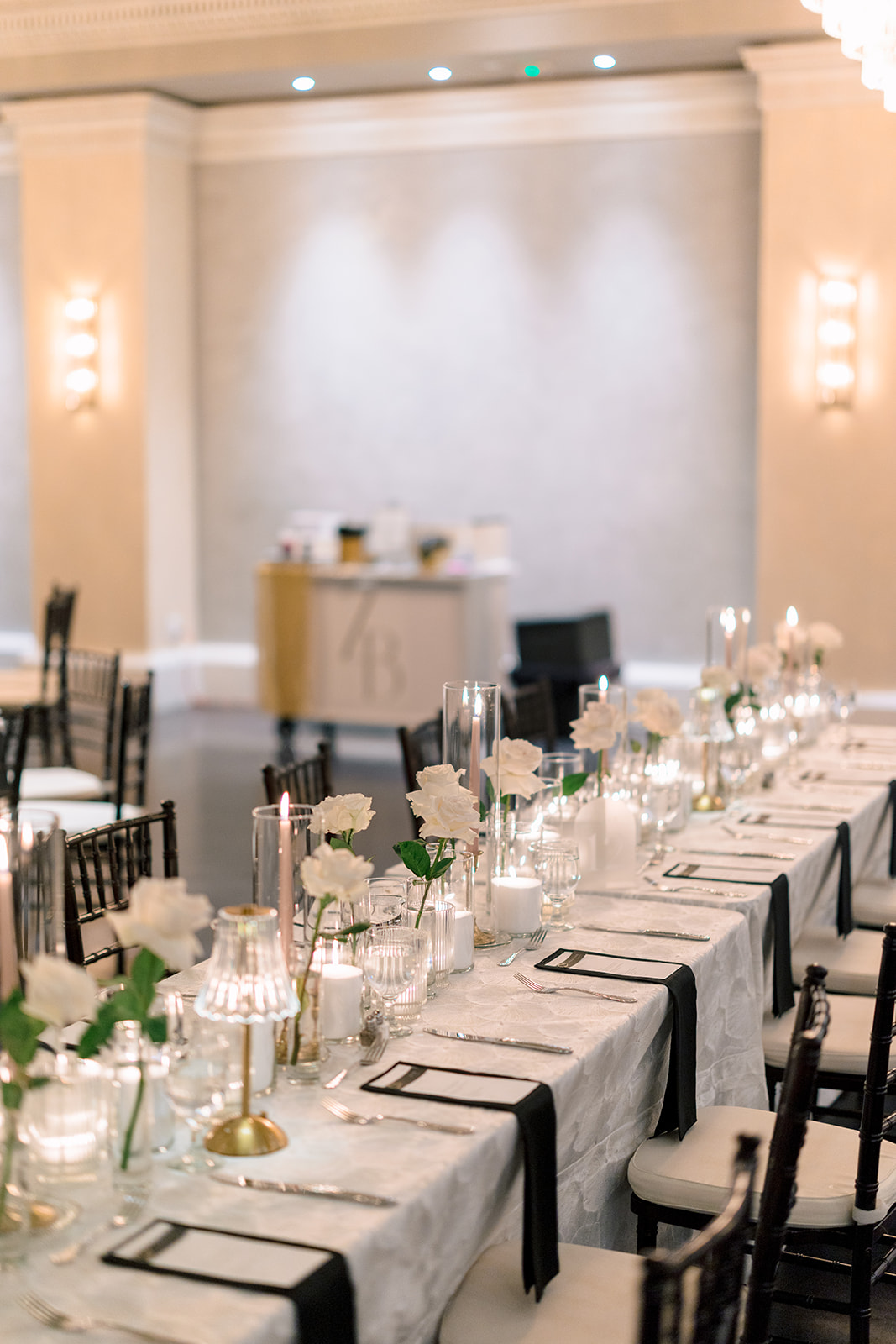A long, elegantly set dining table with white tablecloth, white flowers in vases, candles, glassware, and black napkins in a softly lit room.