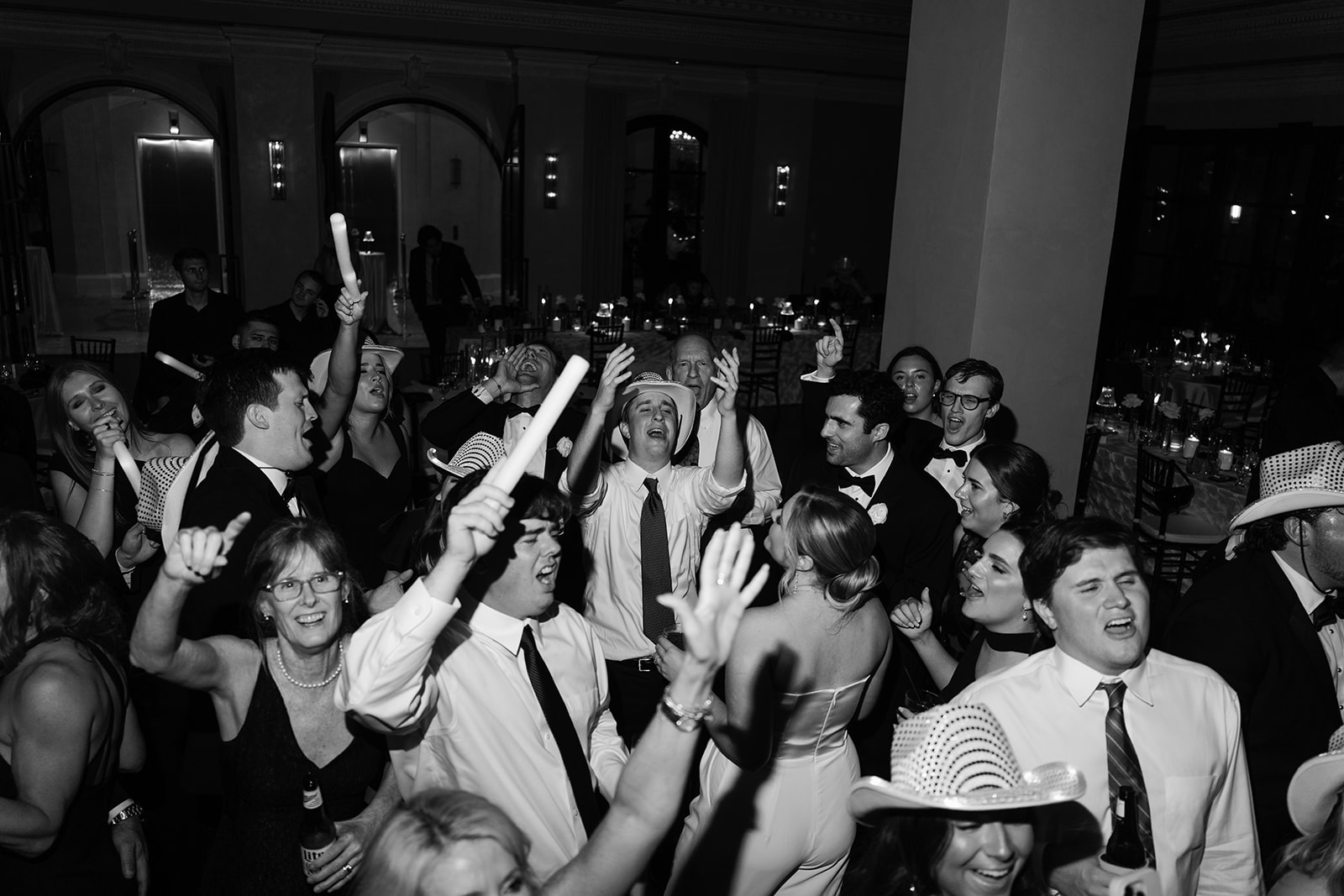 A group of people in formal attire and white hats laugh and dance together at a wedding at Le Tesserae 