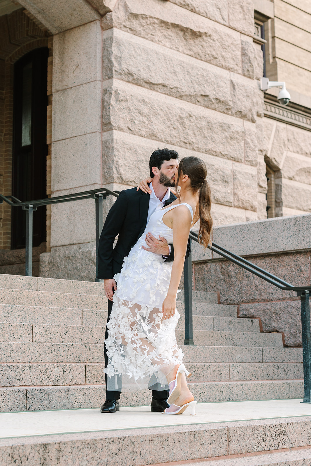 A couple dressed in formal attire stands on stone steps outside a large building; the woman wears a white dress and the man wears a black suit for a  Downtown Houston Engagement Photos 