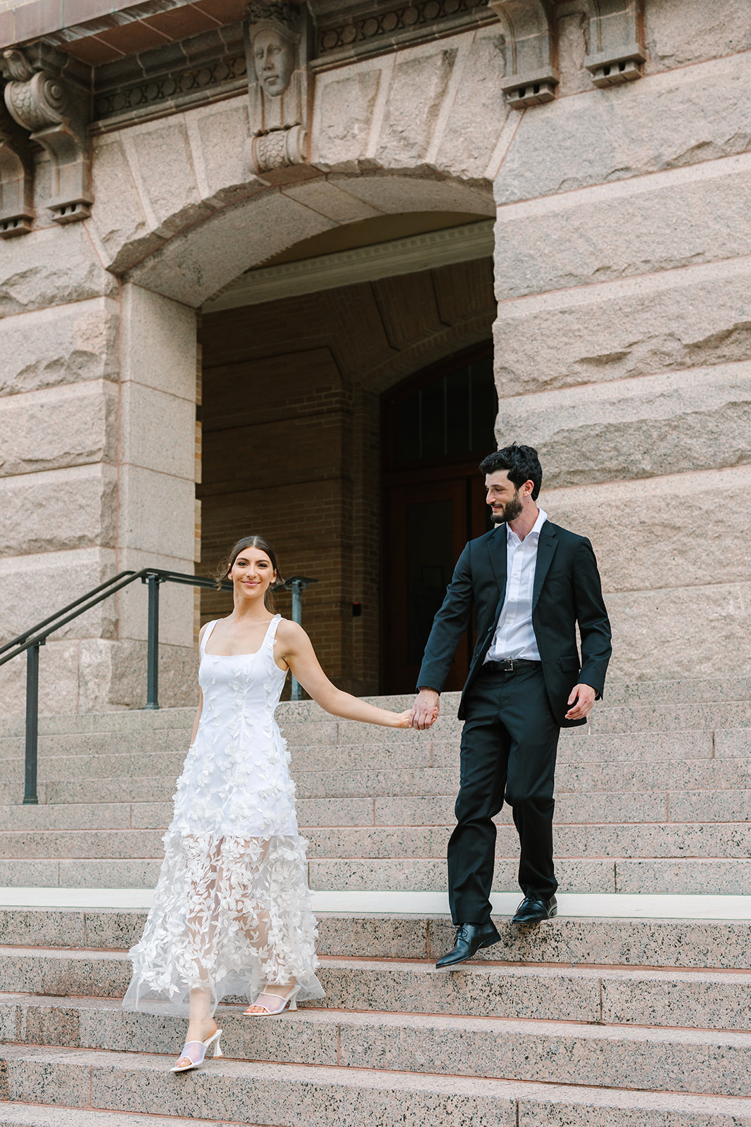 A couple dressed in formal attire stands on stone steps outside a large building; the woman wears a white dress and the man wears a black suit for a  Downtown Houston Engagement Photos 