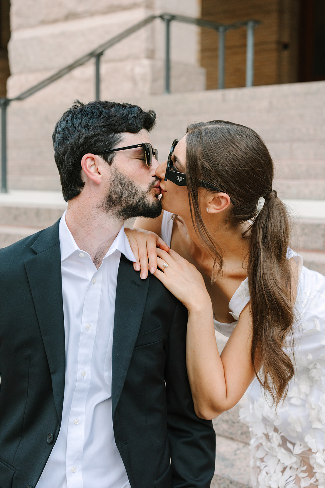 A man in a black suit and a woman in a white dress stand together outdoors on stone steps for their Downtown Houston Engagement Photos 