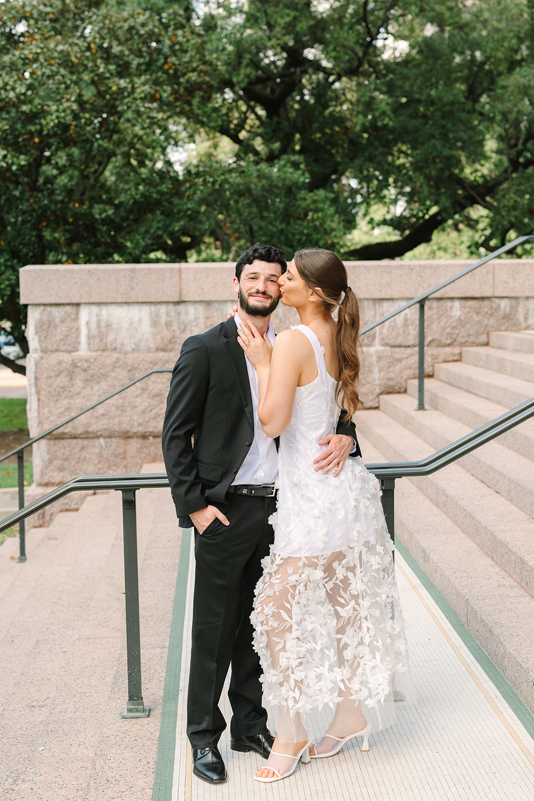 A couple dressed in formal attire stands on stone steps outside a large building; the woman wears a white dress and the man wears a black suit for a  Downtown Houston Engagement Photos 