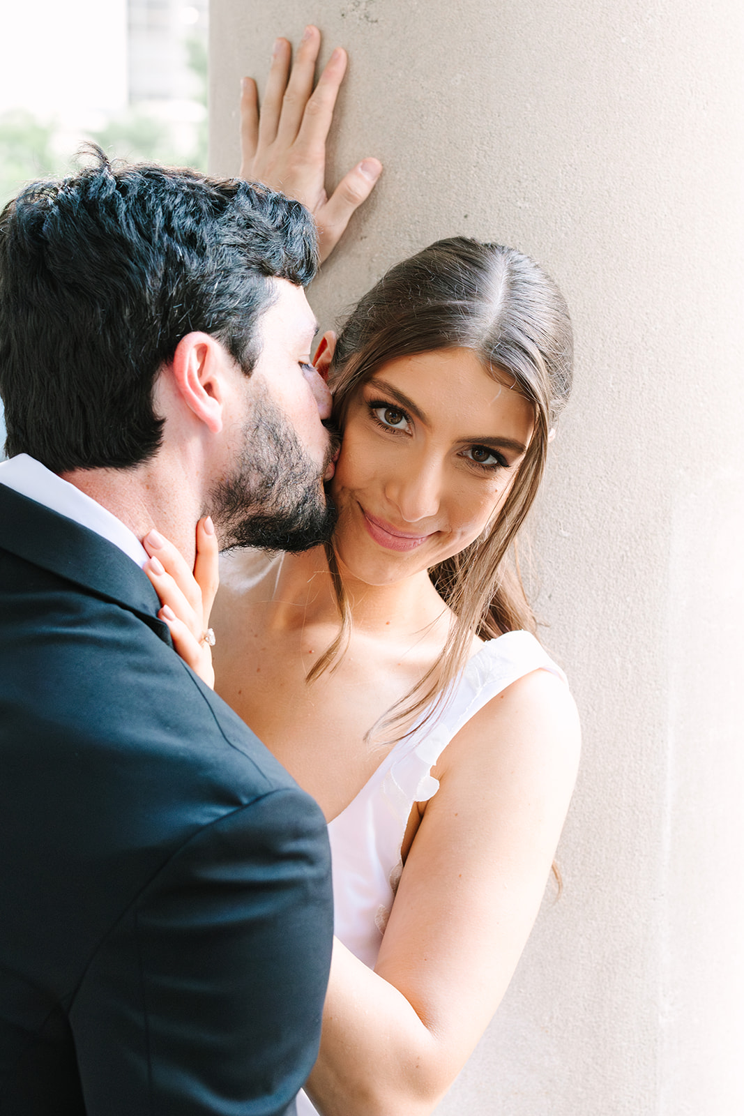 A man in a suit whispers into a woman's ear as she smiles at the camera, with her hand on his shoulder and his hand against a column.
