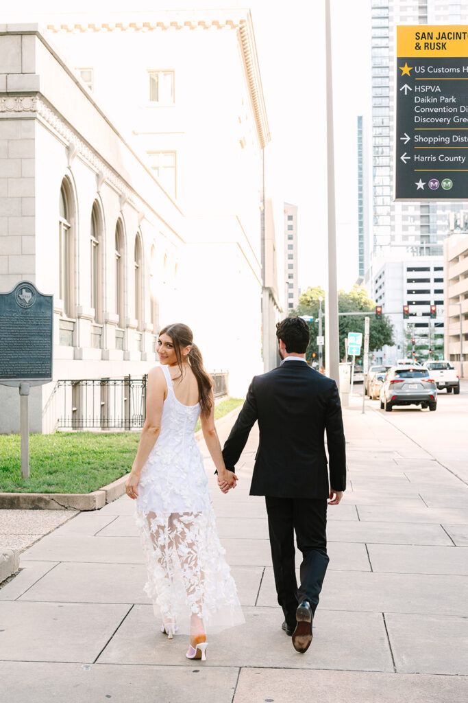 A man in a suit and a woman in a white dress walk hand in hand down a city sidewalk, smiling at each other.