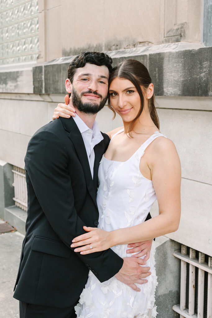 A man in a black suit and a woman in a white sleeveless dress stand close together, smiling and posing for a photo outdoors against a stone building.