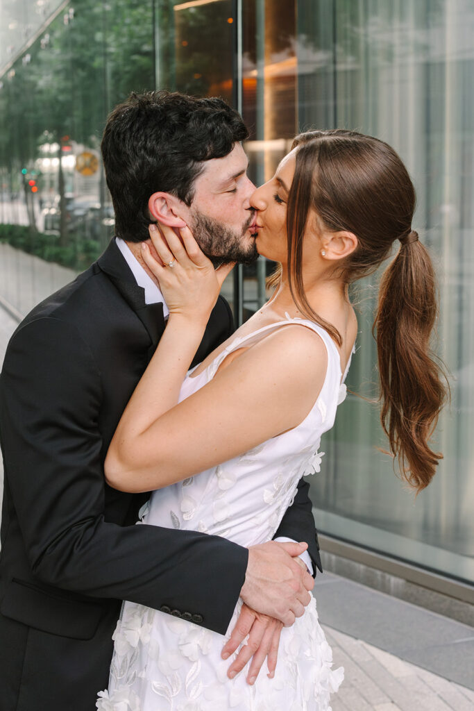 A man in a black suit and a woman in a white sleeveless dress stand close together, smiling and posing for a photo outdoors against a stone building.