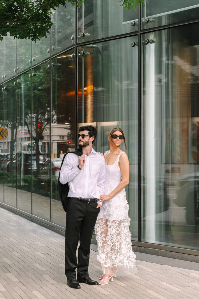 A man in a white shirt and black pants stands next to a woman in a white sleeveless dress, both wearing sunglasses, on a sidewalk by a glass building.