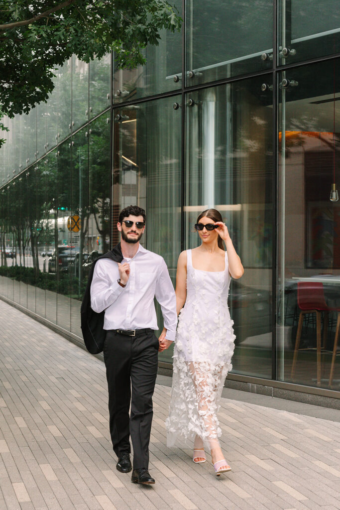 A man in a white shirt and black pants stands next to a woman in a white sleeveless dress, both wearing sunglasses, on a sidewalk by a glass building.