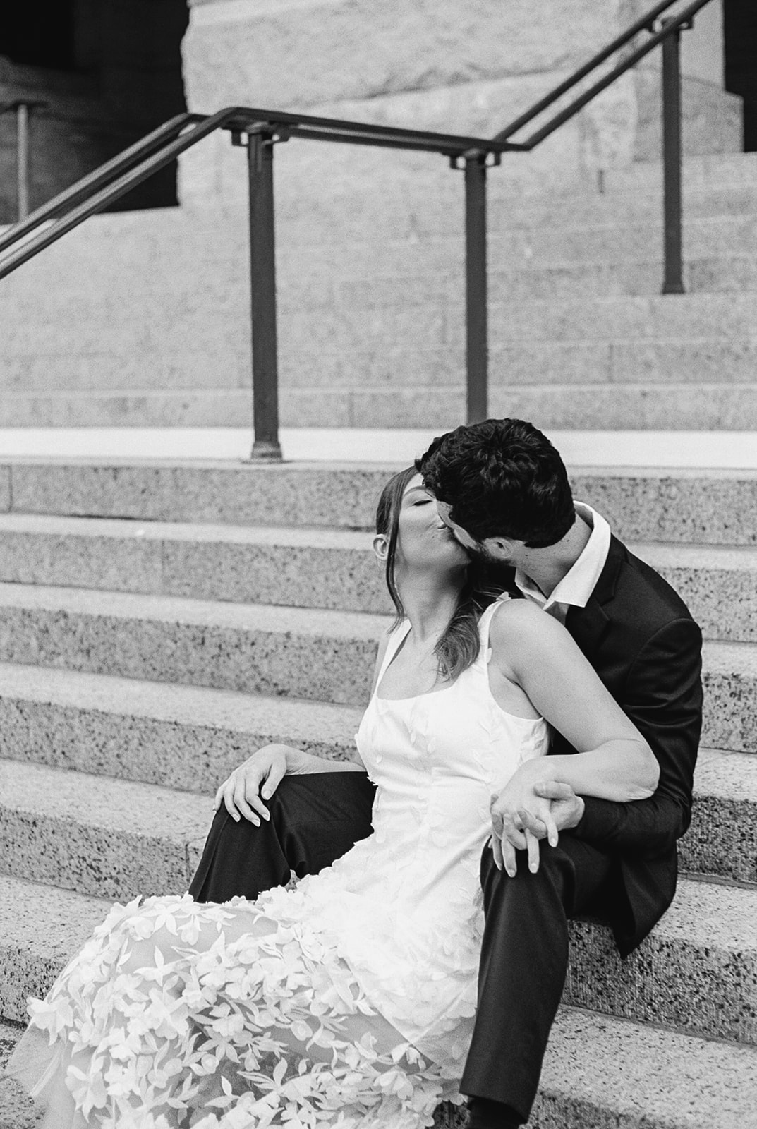 A man and woman, both wearing sunglasses, walk hand in hand and smile. The man holds a jacket over his shoulder. The woman wears a white dress. They are outside in front of a glass building.