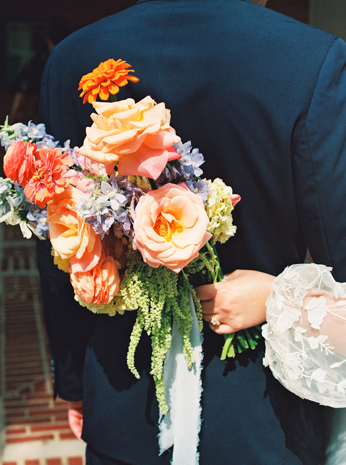 A bride in a white dress and a groom in a dark suit walk hand in hand outdoors, smiling at each other, with trees and brick buildings in the background at a garden wedding venue
