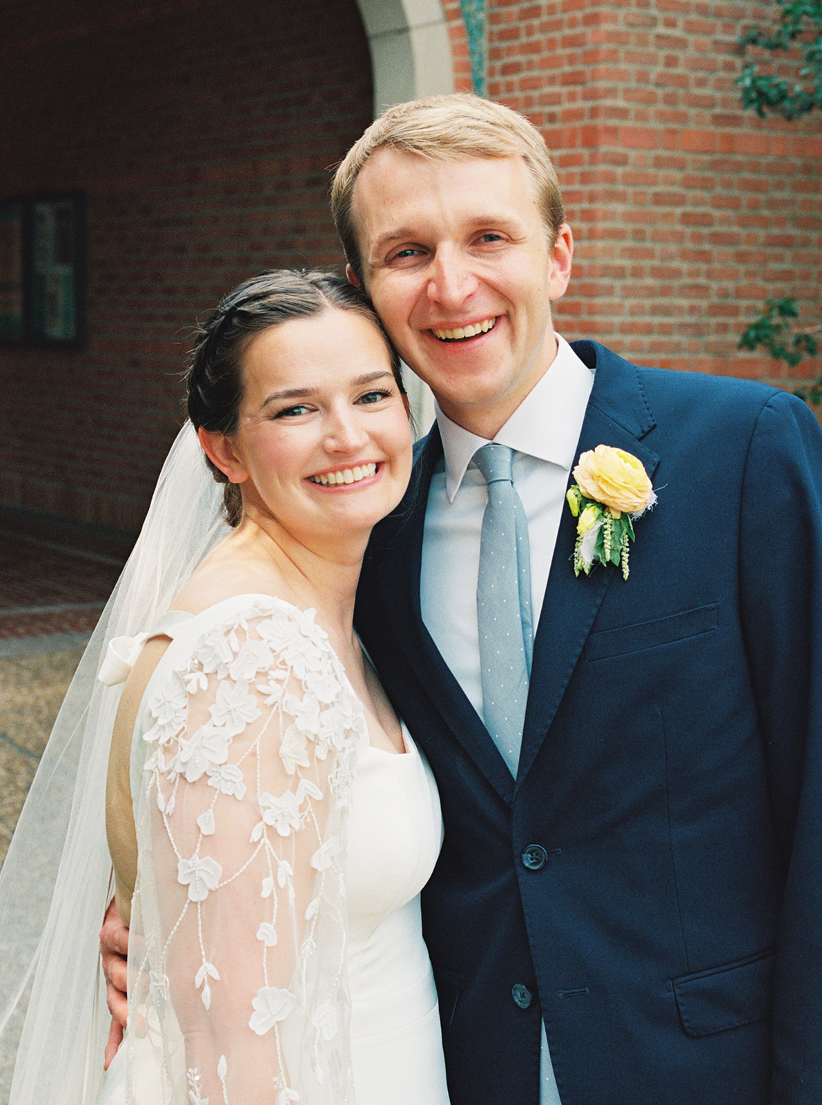 A bride in a white dress and a groom in a dark suit walk hand in hand outdoors, smiling at each other, with trees and brick buildings in the background at a garden wedding venue