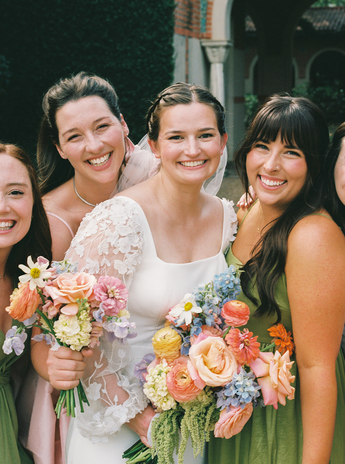 A wedding party poses outdoors in front of a building with arches, with bridesmaids in colorful dresses and groomsmen in dark suits at the garden wedding venue