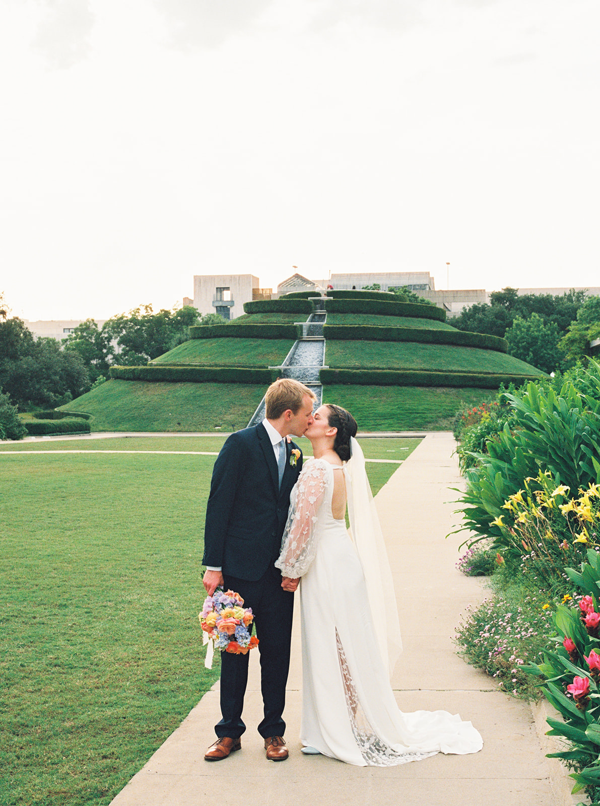 A bride in a white dress and a groom in a dark suit walk hand in hand outdoors, smiling at each other, with trees and brick buildings in the background at a garden wedding venue