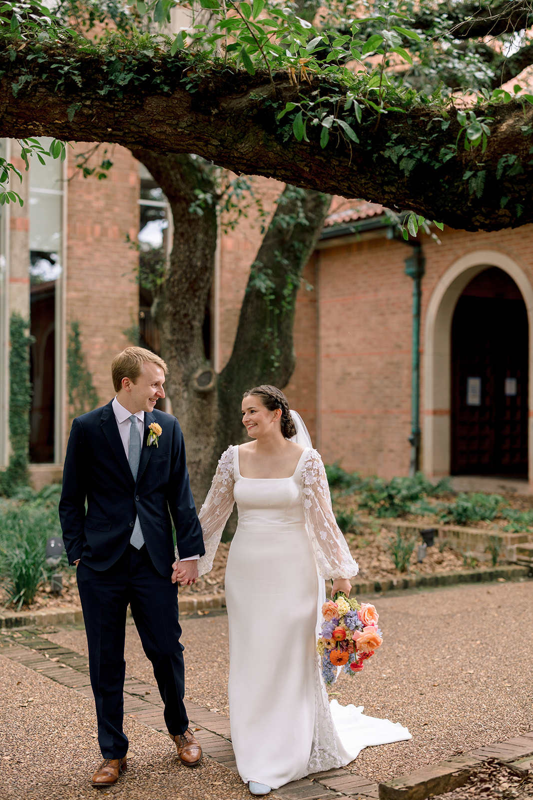 A bride in a white dress and a groom in a dark suit walk hand in hand outdoors, smiling at each other, with trees and brick buildings in the background at a garden wedding venue