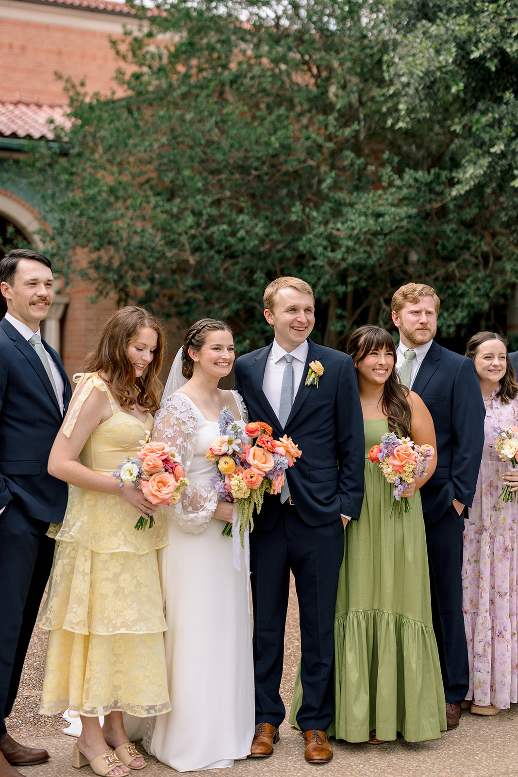A wedding party poses outdoors in front of a building with arches, with bridesmaids in colorful dresses and groomsmen in dark suits at the garden wedding venue