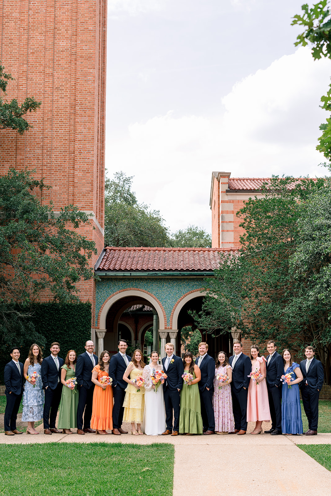 A wedding party poses outdoors in front of a building with arches, with bridesmaids in colorful dresses and groomsmen in dark suits at the garden wedding venue