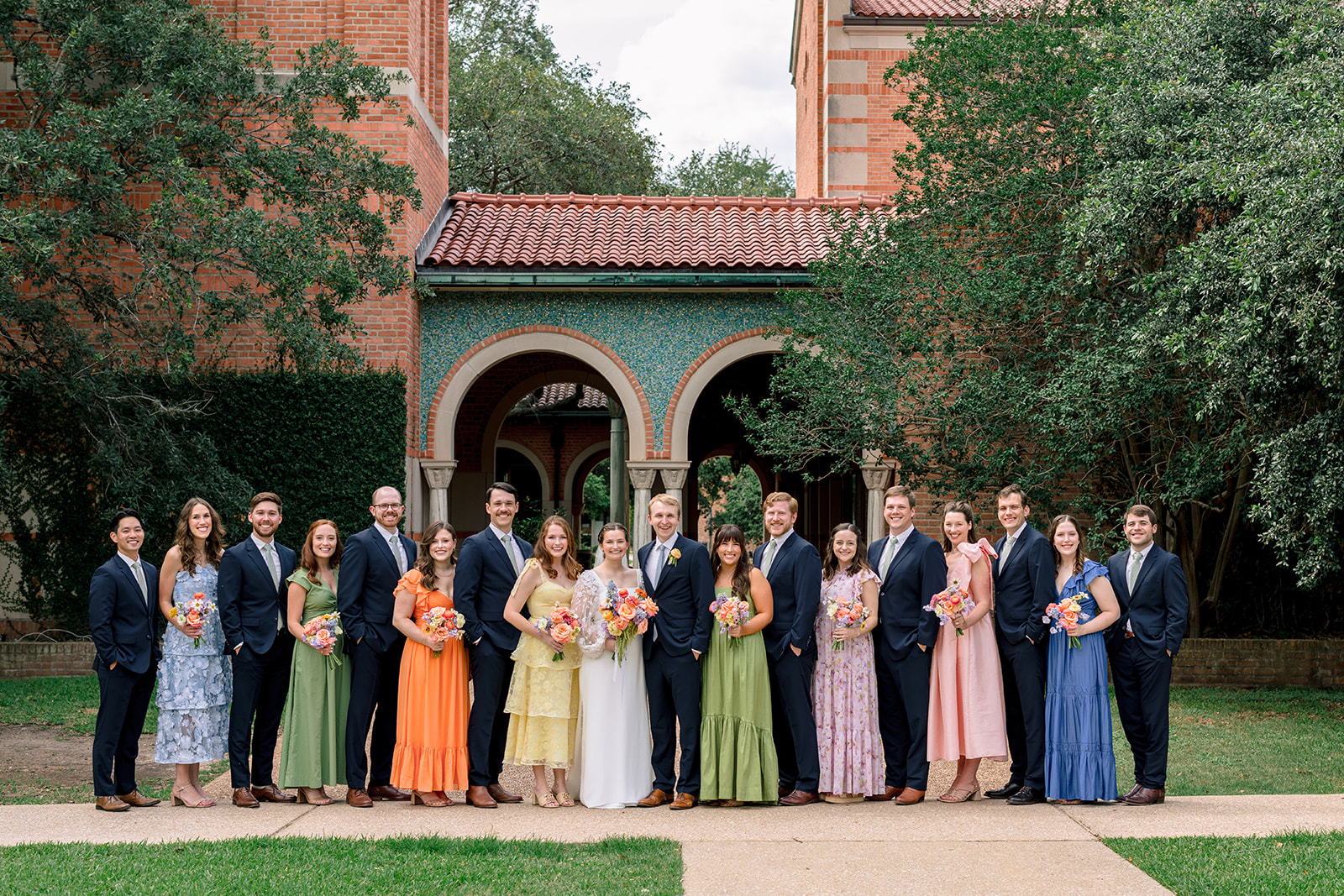 A wedding party poses outdoors in front of a building with arches, with bridesmaids in colorful dresses and groomsmen in dark suits at the garden wedding venue