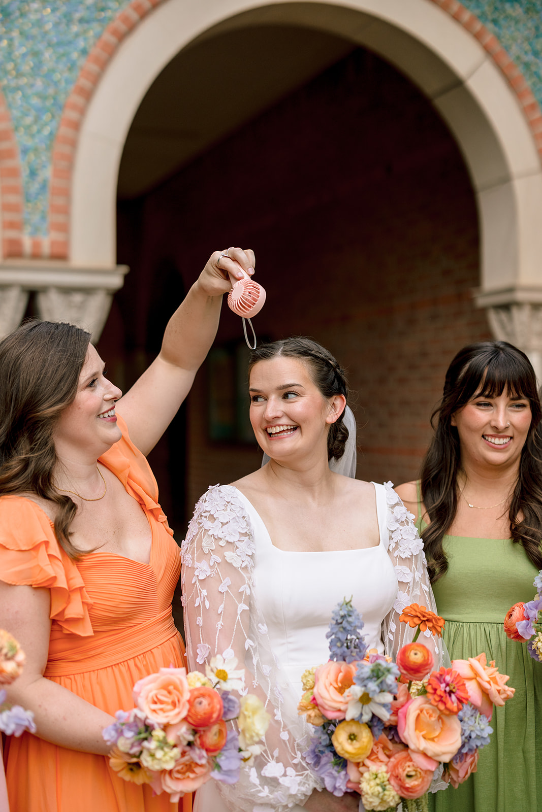 A wedding party poses outdoors in front of a building with arches, with bridesmaids in colorful dresses and groomsmen in dark suits at the garden wedding venue