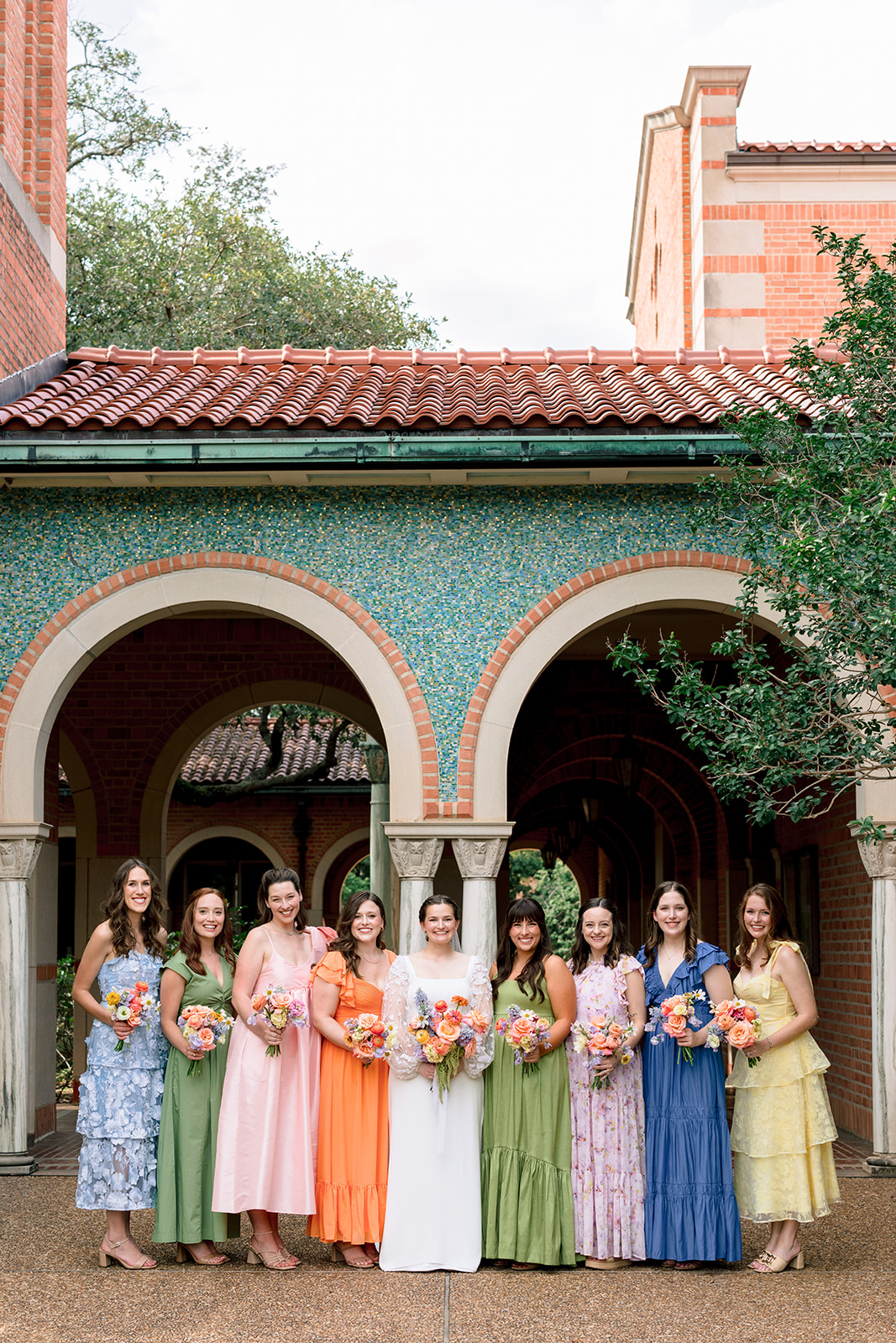 A wedding party poses outdoors in front of a building with arches, with bridesmaids in colorful dresses and groomsmen in dark suits at the garden wedding venue