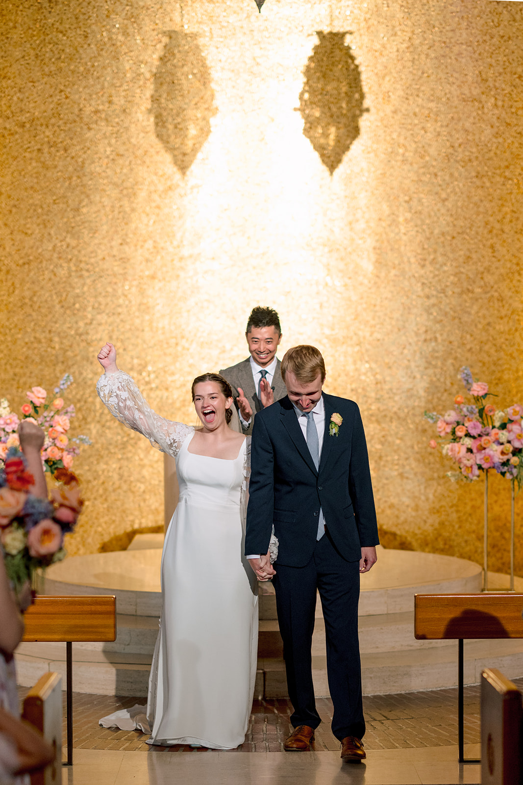 A bride and groom stand at the altar with an officiant in a church filled with golden light, as guests watch the wedding ceremony from the pews.