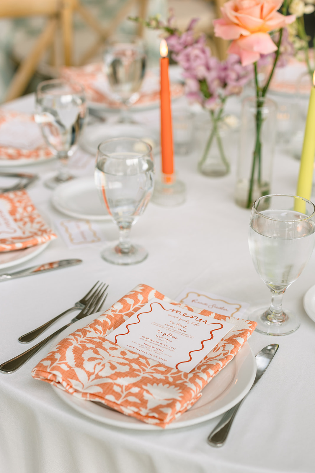 A long, elegantly set dining table is decorated with colorful flowers in vases, glassware, and plates, situated in a bright room with large windows at a garden wedding venue